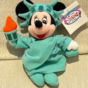 May include: A plush toy of Mickey Mouse dressed as the Statue of Liberty. The toy is holding a torch in its right hand. The toy is wearing a green dress and a crown with spikes. The toy has a tag that says "The Disney Store".