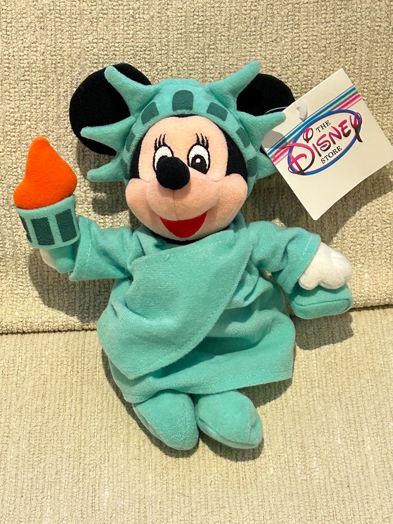 May include: A plush toy of Mickey Mouse dressed as the Statue of Liberty. The toy is holding a torch in its right hand. The toy is wearing a green dress and a crown with spikes. The toy has a tag that says "The Disney Store".