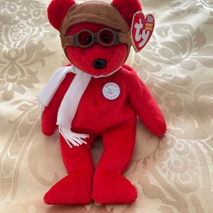 Puede incluir: Un oso de peluche rojo con gorro y gafas de aviador marrones, una bufanda blanca y una etiqueta. El oso tiene una nariz negra y un parche circular que dice "100 Years of Flight".