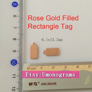 May include: Two rose gold filled rectangle tags with a hole at the top for hanging. The tags measure 6.1 mm by 13.7 mm. The tags are on a white surface with a ruler for size reference. The text "Etsy: Umonograms" is visible in the image.