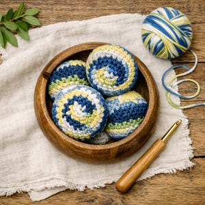 May include: A round wooden bowl containing four crocheted spheres with a spiral pattern in shades of blue, yellow, and white. Beside the bowl sits a ball of matching variegated yarn and a wooden crochet hook with a gold-colored metal tip.