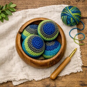 May include: A round wooden bowl containing four crocheted spheres in shades of blue, green, and gray. Beside the bowl sits a ball of variegated blue and green yarn and a wooden crochet hook with a gold-colored metal tip and a smooth handle.