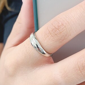 May include: A polished silver ring with a flowing, curved design. The ring is worn on a finger, reflecting light. This piece of jewellery is a simple, elegant accessory.