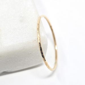 Peut inclure: Une fine bague en métal de couleur dorée avec une surface martelée et texturée. L'anneau est circulaire et présente une finition réfléchissante qui capte la lumière, créant de petites facettes distinctes sur le bord extérieur du bijou.