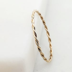 May include: A delicate gold-colored ring with a twisted rope design. The ring is thin and sits on a white surface. The ring's design is a continuous twist, creating a textured appearance. The ring is a simple, elegant piece of jewelry.