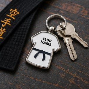 May include: A silver-toned metal keyring shaped like a martial arts uniform top. The white enamel centre features a black belt graphic and the text CLUB NAME in black lettering. A silver split ring holds two metal keys attached to the keyring.
