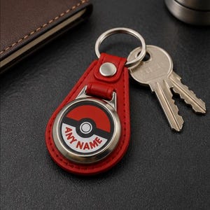 May include: A red leather keyring holder featuring a circular metal insert with a red and white graphic design resembling a ball, including a central black band and the text ANY NAME printed in red capital letters, attached to a silver ring.