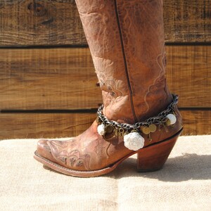 May include: A brown leather cowboy boot with decorative stitching and a pointed toe. The boot features a decorative chain with charms and white floral accents. The boot is set against a rustic wooden background.