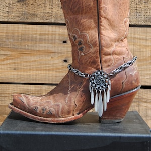 May include: Brown leather cowboy boot with a silver chain and a decorative pendant with a rhinestone flower. The boot is on a black surface.