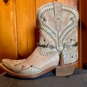 May include: A beige leather cowboy boot with decorative stitching, metal studs, and a chain ankle bracelet with a tassel. The boot has a pointed toe and a stacked heel.