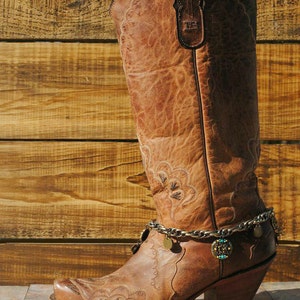 May include: A brown leather cowboy boot with intricate stitching and a decorative chain ankle bracelet. The boot has a pointed toe and a stacked heel. The background is a wooden wall.