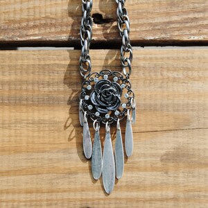May include: A silver chain necklace with a black rose pendant and silver teardrop-shaped charms. The pendant is adorned with clear rhinestones.