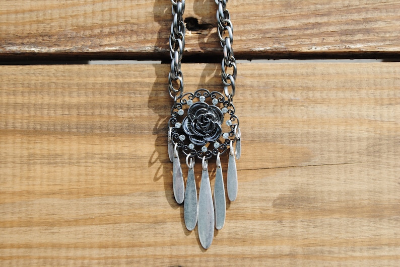 May include: A silver chain necklace with a black rose pendant and silver teardrop-shaped charms. The pendant is adorned with clear rhinestones.