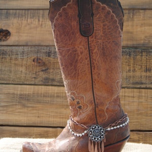 May include: A brown leather cowboy boot with decorative stitching and a pointed toe. The boot features a decorative chain and a floral embellishment with dangling accents. The boot is set against a wooden background.