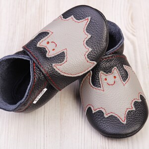 May include: A pair of black leather baby shoes with a gray bat applique. The bat has red stitching and a smiling face. The shoes are designed for infants and toddlers.