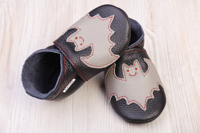 May include: A pair of black leather baby shoes with a gray bat applique. The bat has red stitching and a smiling face. The shoes are designed for infants and toddlers.