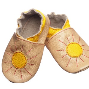 May include: A pair of beige leather baby shoes with a yellow sun design. The sun has red stitching around it.