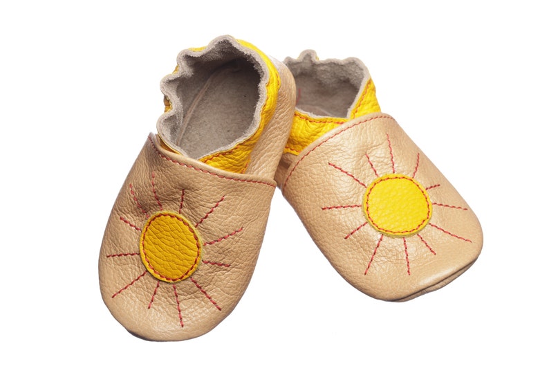 May include: A pair of beige leather baby shoes with a yellow sun design. The sun has red stitching around it.