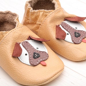 May include: A pair of tan leather baby shoes with a dog face design. The shoes have brown ears, a white face, a brown nose, and a pink tongue.