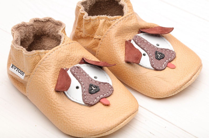 May include: A pair of tan leather baby shoes with a dog face design. The shoes have brown ears, a white face, a brown nose, and a pink tongue.