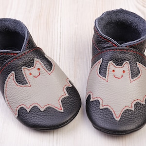 May include: A pair of black leather baby shoes with a gray bat applique stitched with red thread. The shoes have a soft sole and a dark gray interior. The brand name is visible on the side of one shoe.