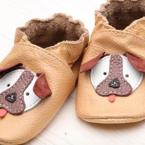 May include: A pair of tan leather baby shoes with a dog face design. The shoes have brown ears, a white nose, and a red tongue.