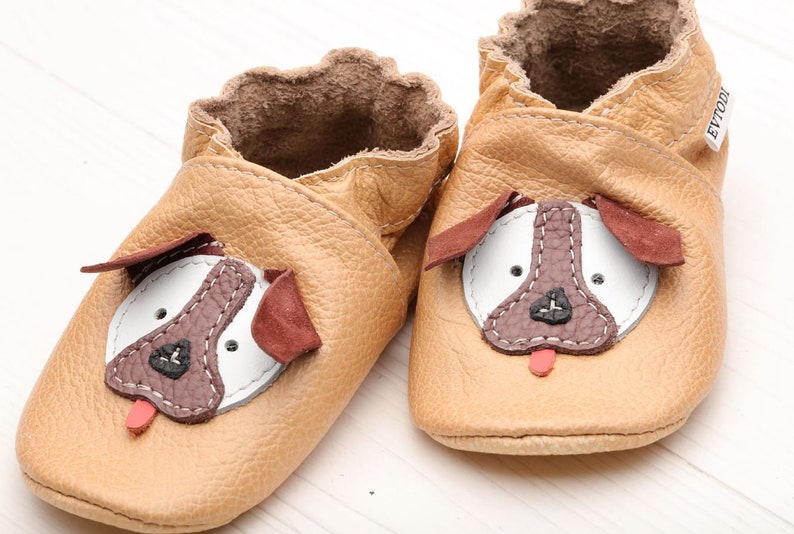 May include: A pair of tan leather baby shoes with a dog face design. The shoes have brown ears, a white nose, and a red tongue.