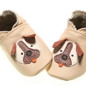 May include: A pair of beige leather baby shoes with a dog face design. The shoes have brown ears, a brown nose, and white eyes.