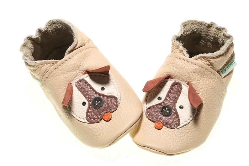 May include: A pair of beige leather baby shoes with a dog face design. The shoes have brown ears, a brown nose, and white eyes.
