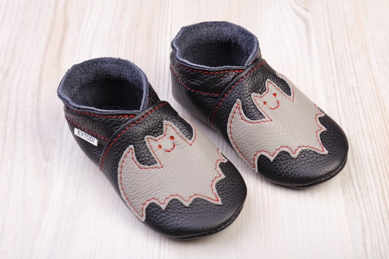 May include: A pair of black leather baby shoes with a gray bat applique and red stitching. The shoes have a soft sole and a dark blue interior. The brand name "EVTODI" is visible on one shoe.