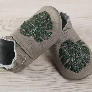 May include: A pair of grey leather baby shoes with green monstera leaf embroidery.