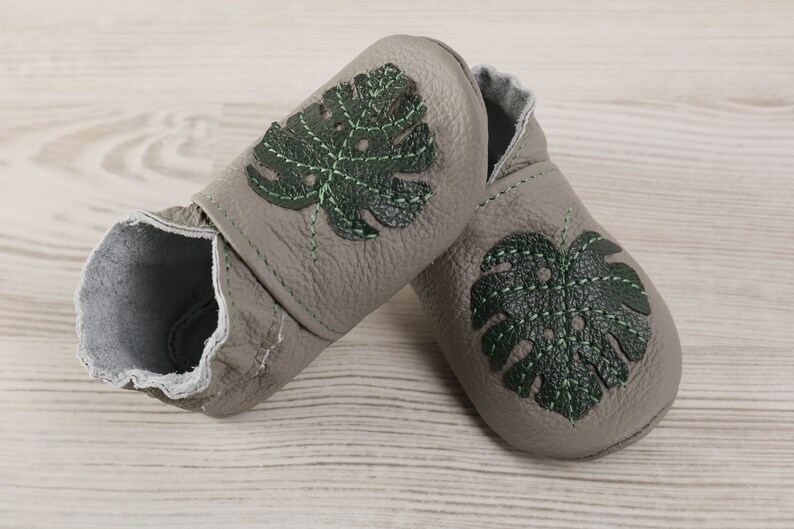 May include: A pair of grey leather baby shoes with green monstera leaf embroidery.
