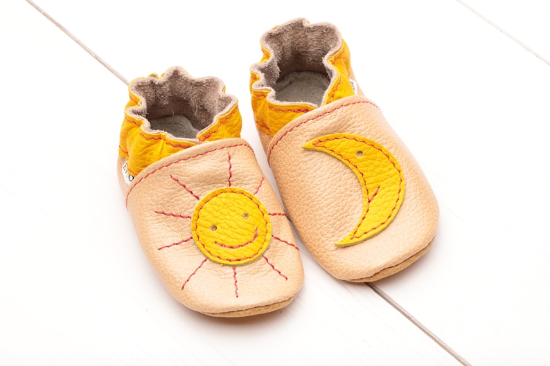 May include: A pair of baby shoes with a sun and moon design. The shoes are made of soft leather and have a yellow and beige color scheme.