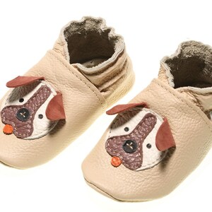 May include: A pair of beige leather baby shoes with a dog face design. The shoes have a brown nose, white eyes, and red ears. The shoes are made by the brand Evtodi.