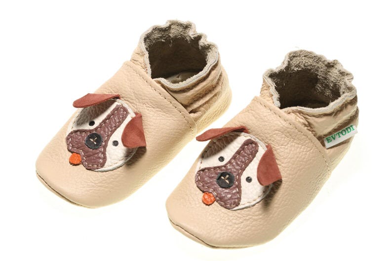 May include: A pair of beige leather baby shoes with a dog face design. The shoes have a brown nose, white eyes, and red ears. The shoes are made by the brand Evtodi.
