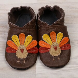 Puede incluir: Un par de zapatos de beb&eacute; de cuero marr&oacute;n con un dise&ntilde;o de pavo. El pavo tiene una cola en forma de abanico roja, naranja y amarilla y un cuerpo marr&oacute;n con un pico rojo.