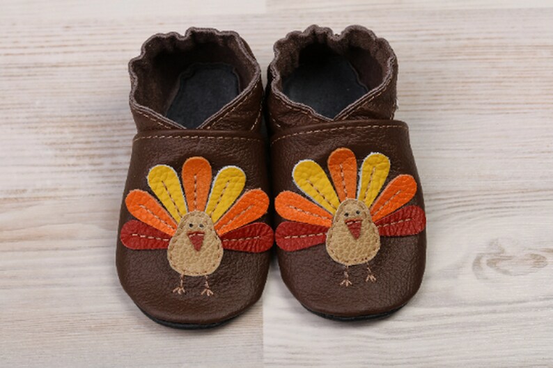 Puede incluir: Un par de zapatos de beb&eacute; de cuero marr&oacute;n con un dise&ntilde;o de pavo. El pavo tiene una cola en forma de abanico roja, naranja y amarilla y un cuerpo marr&oacute;n con un pico rojo.