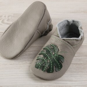 May include: A pair of grey leather baby shoes with green embroidered monstera leaves. The shoes have a soft, flexible sole and are perfect for little feet.