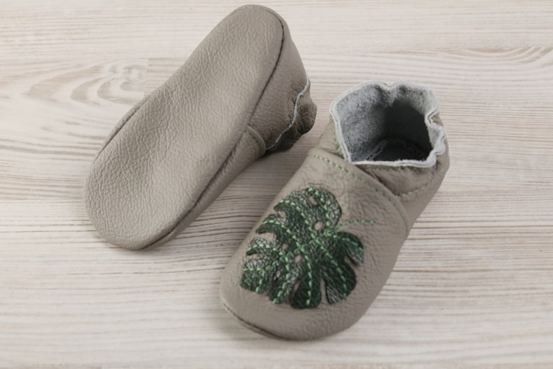 May include: A pair of grey leather baby shoes with green embroidered monstera leaves. The shoes have a soft, flexible sole and are perfect for little feet.