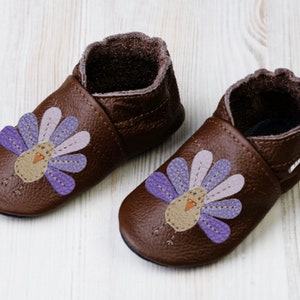 Puede incluir: Un par de zapatos de beb&eacute; de cuero marr&oacute;n con un dise&ntilde;o de pavo morado y rosa. Los zapatos tienen una suela suave y flexible y un ajuste c&oacute;modo.