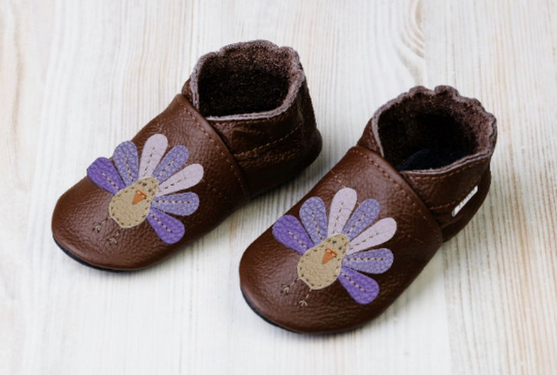 Puede incluir: Un par de zapatos de beb&eacute; de cuero marr&oacute;n con un dise&ntilde;o de pavo morado y rosa. Los zapatos tienen una suela suave y flexible y un ajuste c&oacute;modo.