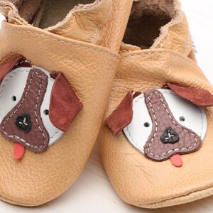 May include: A pair of tan leather baby shoes with a dog face design. The dog face features brown, white, and red accents.