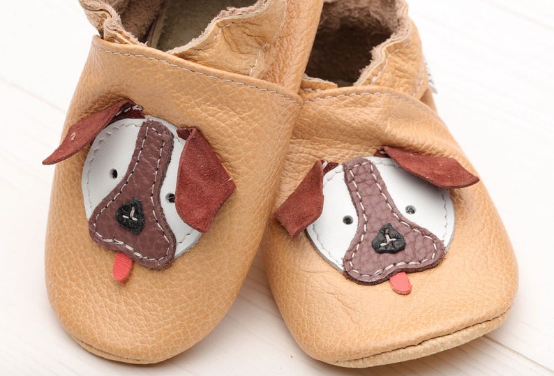 May include: A pair of tan leather baby shoes with a dog face design. The dog face features brown, white, and red accents.