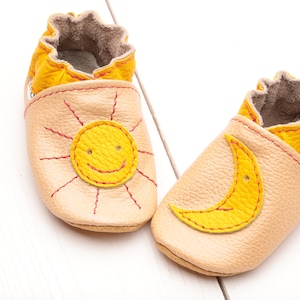 May include: A pair of baby shoes with a sun and moon design. The shoes are made of soft leather and have a yellow and beige color scheme. The sun has a smiling face and the moon is a crescent shape.