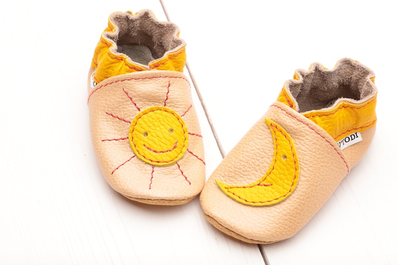 May include: A pair of baby shoes with a sun and moon design. The shoes are made of soft leather and have a yellow and beige color scheme. The sun has a smiling face and the moon is a crescent shape.
