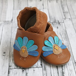 Puede incluir: Un par de zapatos de beb&eacute; de cuero marr&oacute;n con un dise&ntilde;o de pavo. El pavo tiene plumas de cola azules, azul verdoso y marrones y un pico amarillo.