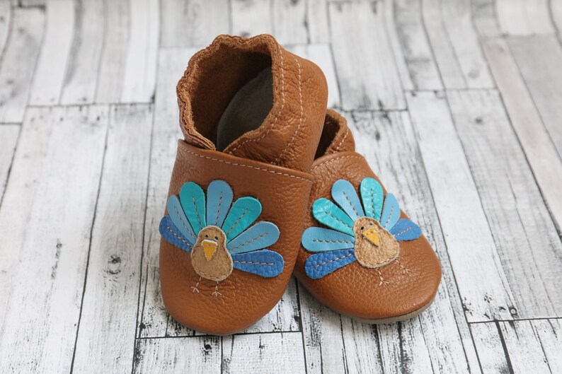 Puede incluir: Un par de zapatos de beb&eacute; de cuero marr&oacute;n con un dise&ntilde;o de pavo. El pavo tiene plumas de cola azules, azul verdoso y marrones y un pico amarillo.