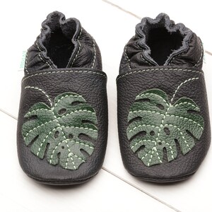 May include: A pair of black leather baby shoes with green monstera leaf cutouts. The shoes have a soft, flexible sole and are ideal for little feet.