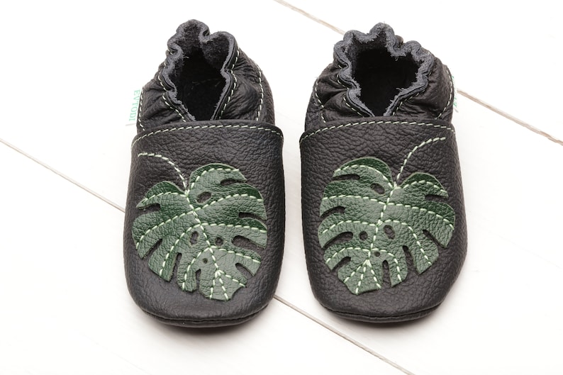 May include: A pair of black leather baby shoes with green monstera leaf cutouts. The shoes have a soft, flexible sole and are ideal for little feet.