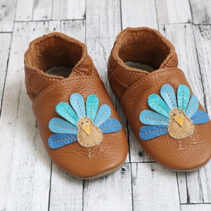 Puede incluir: Un par de zapatos de beb&eacute; de cuero marr&oacute;n con un dise&ntilde;o de pavo. El pavo tiene un dise&ntilde;o de plumas azules, verde azulado y amarillas.